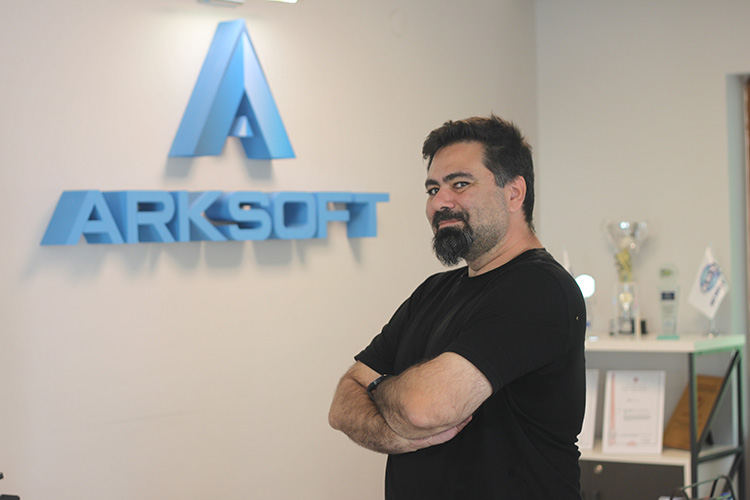 Direnç Önen, CTO of ARKSOFT, in a casual stance with arms crossed, in front of the company logo wall